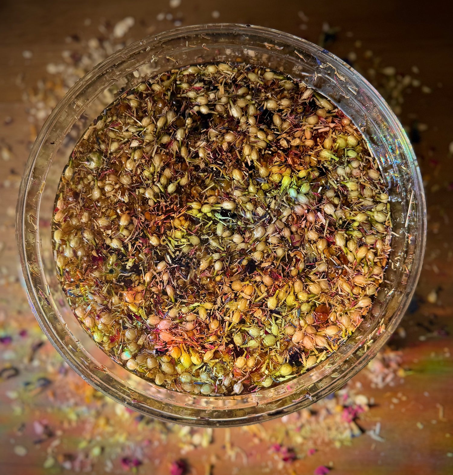 Clear bowl filled with colorful dried herbs, flowers, and oil on a wooden background